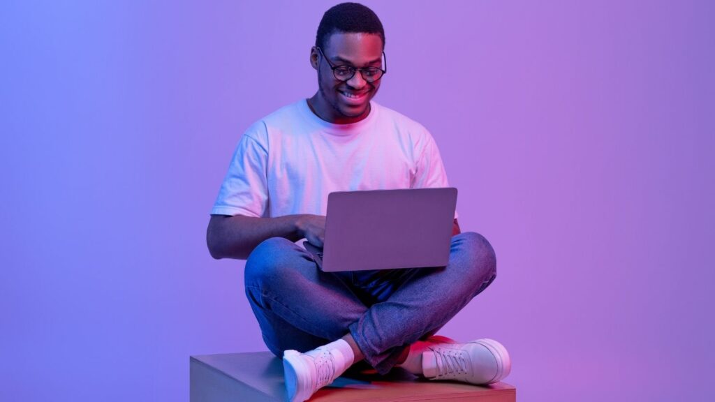 Programmer Guy Using Laptop Computer, Sitting On Cube In Neon Lighting
